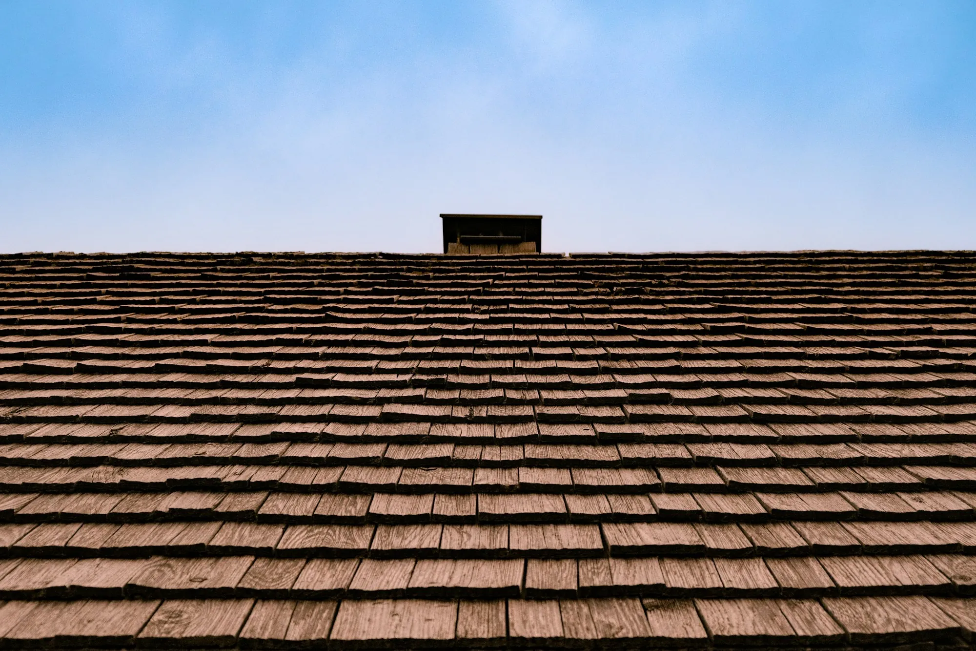 Weathered wood shingle roof with a small chimney cap against blue sky